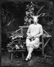 Una donna seduta su una panchina in un giardino, sulla testa una maschera-testa di un animale con due corna.